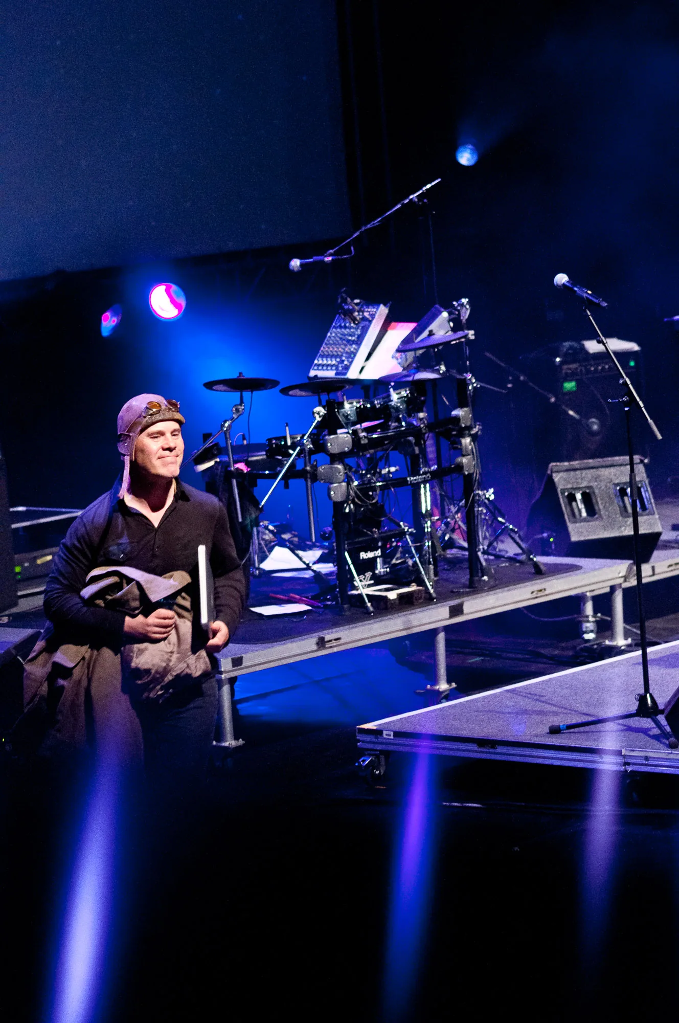 Backstage crew member in an aviator flying cap holding gear near an electronic drum kit under blue stage lighting, live event photography by Hector's Eye