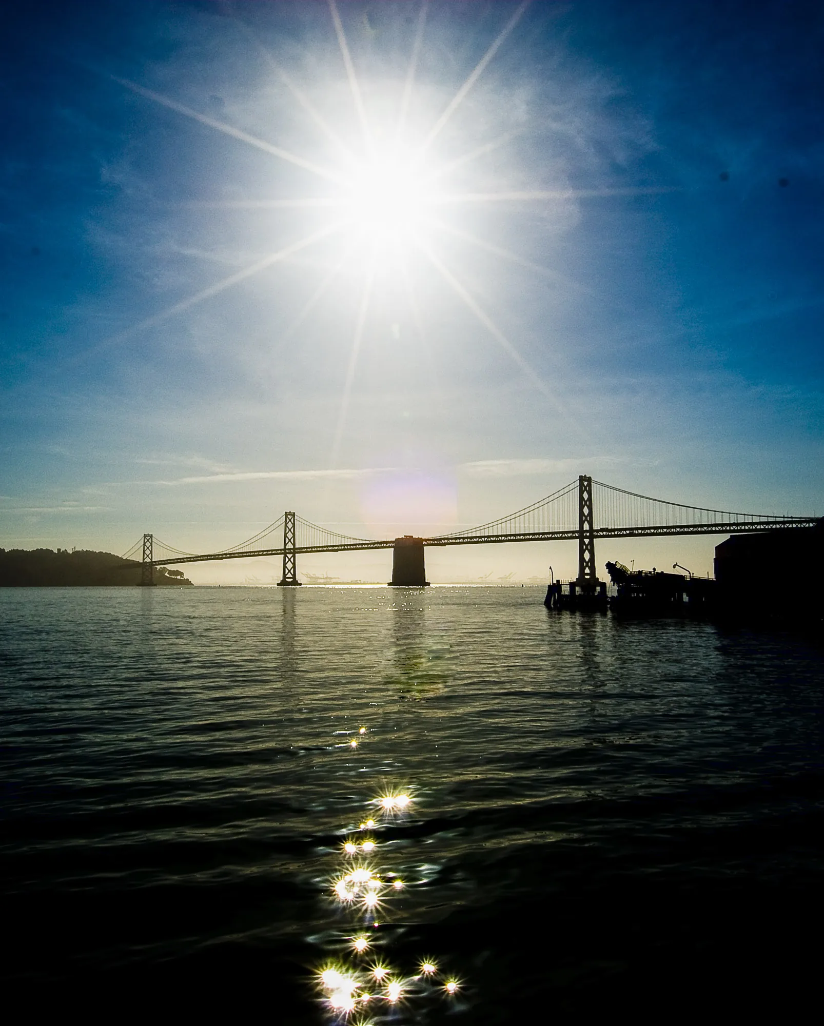 Suspension bridge silhouetted against a bright sunburst over sparkling water, landscape photography
