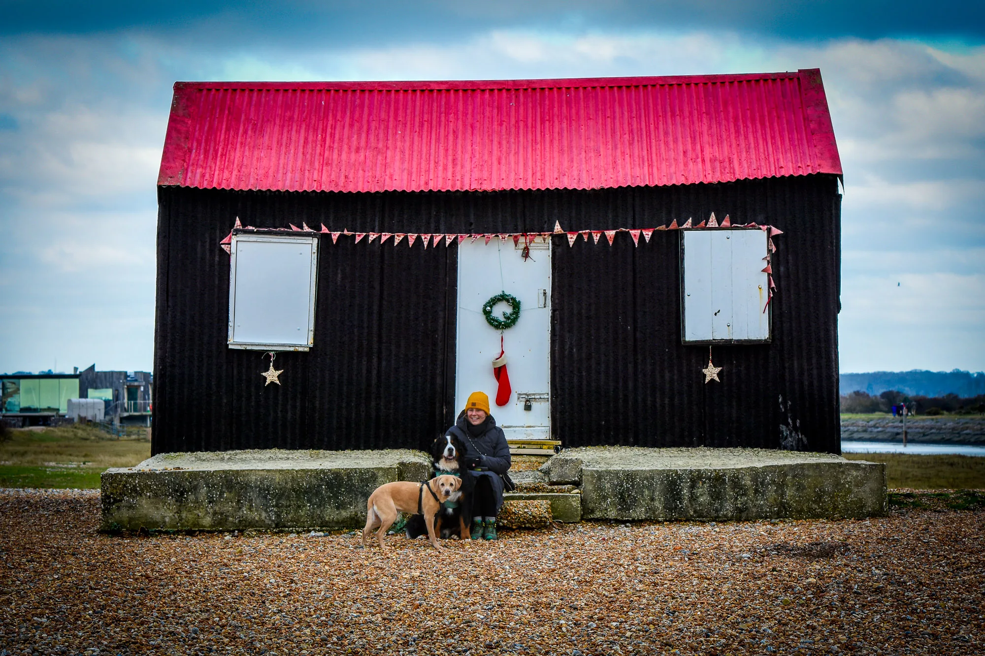 Woman sitting with two dogs outside a Christmas-decorated black beach hut with a red roof, personal photography by Hector's Eye