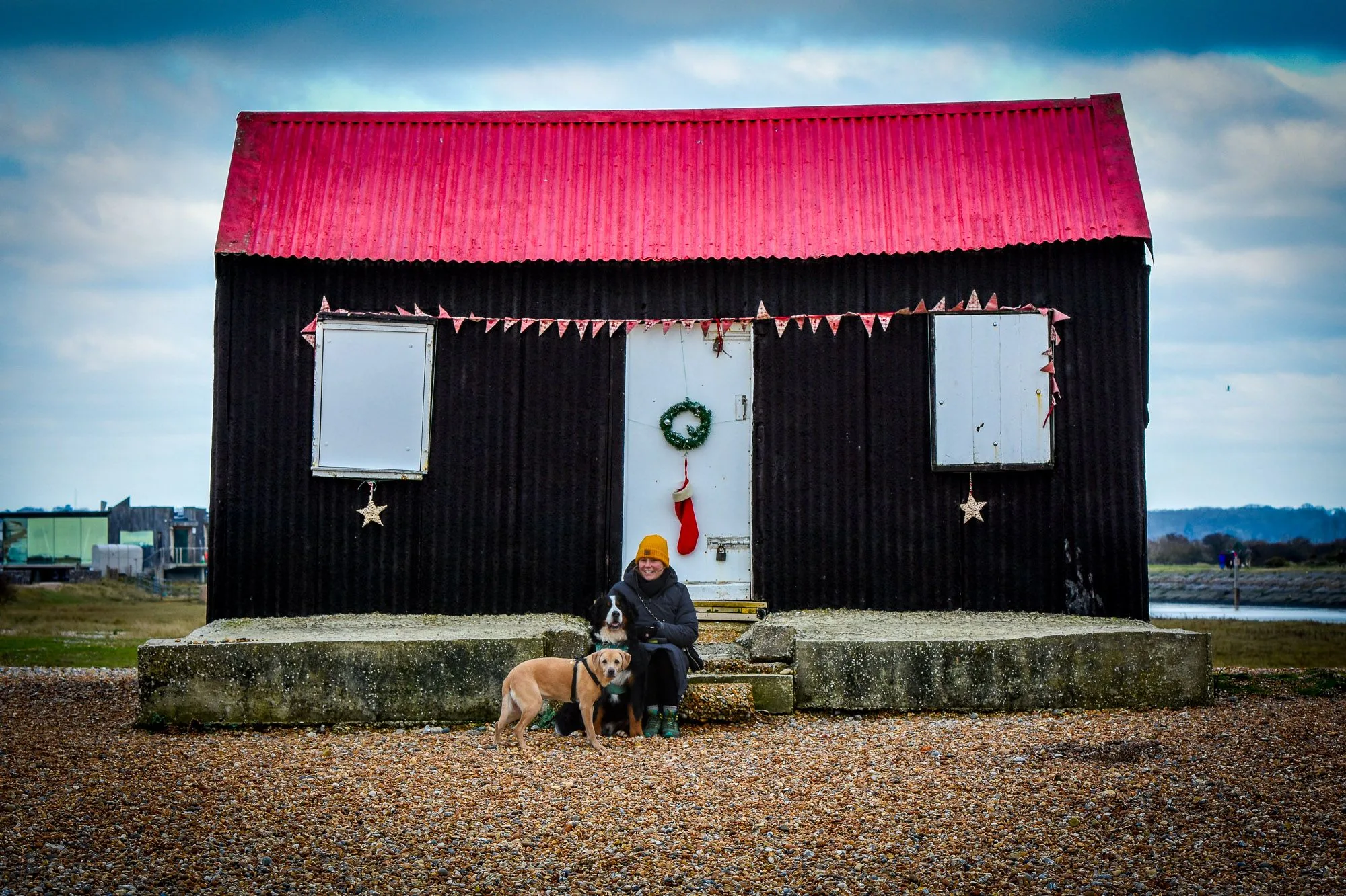 Person and dog sitting outside a red-roofed beach hut on the coast, personal photography by Hector's Eye