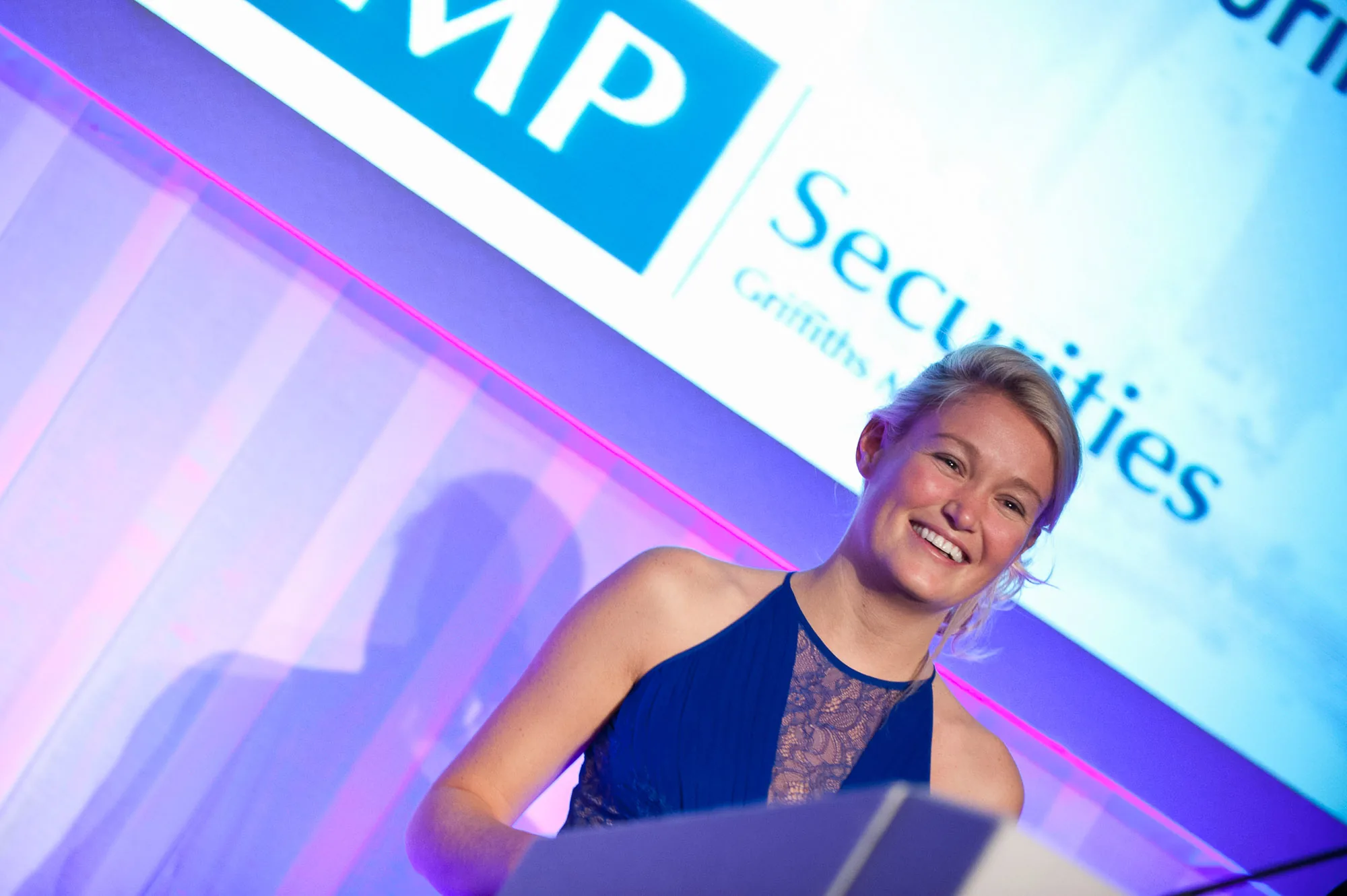 Smiling speaker on stage lit by pink and blue lighting at a conference, corporate event photography by Hector's Eye