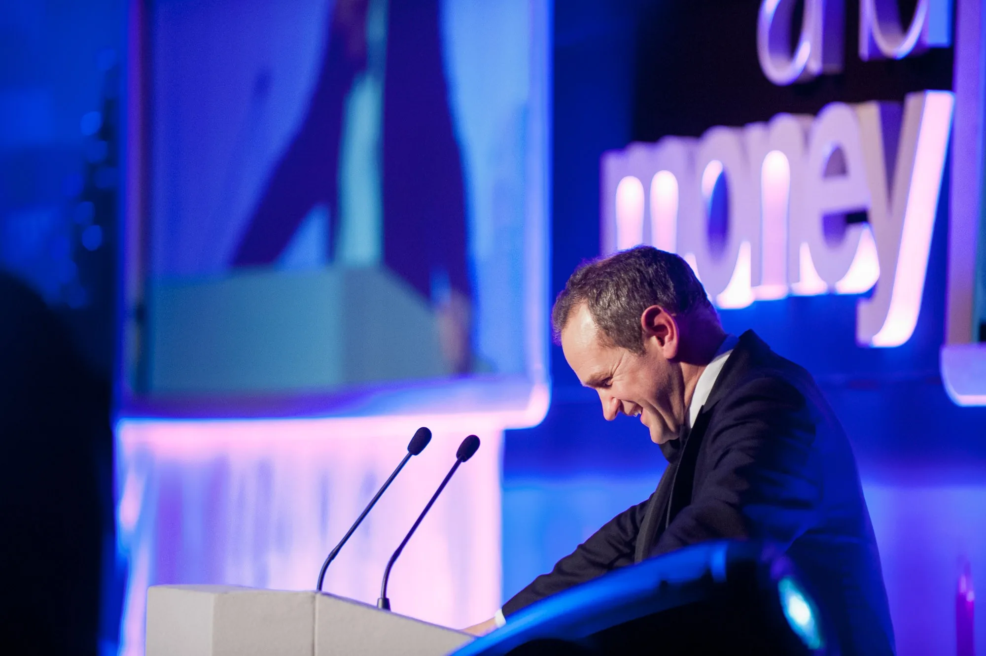 Host smiling at a lectern in front of illuminated event signage, corporate awards photography by Hector's Eye