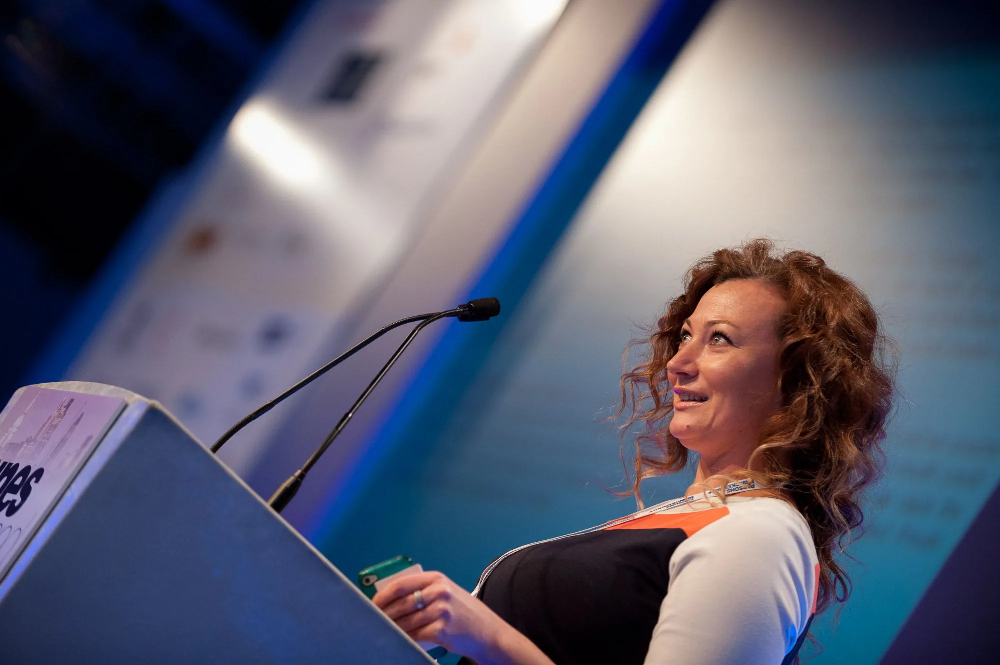 Female speaker addressing an audience from a podium under blue conference lighting, event photography by Hector's Eye