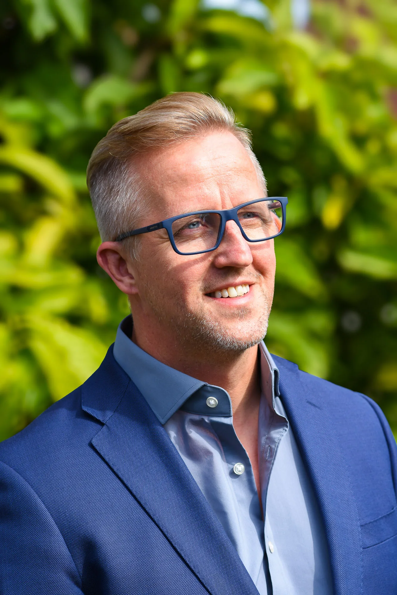 Smiling corporate headshot of a man wearing glasses and a navy blazer outdoors, corporate event photography by Hector's Eye