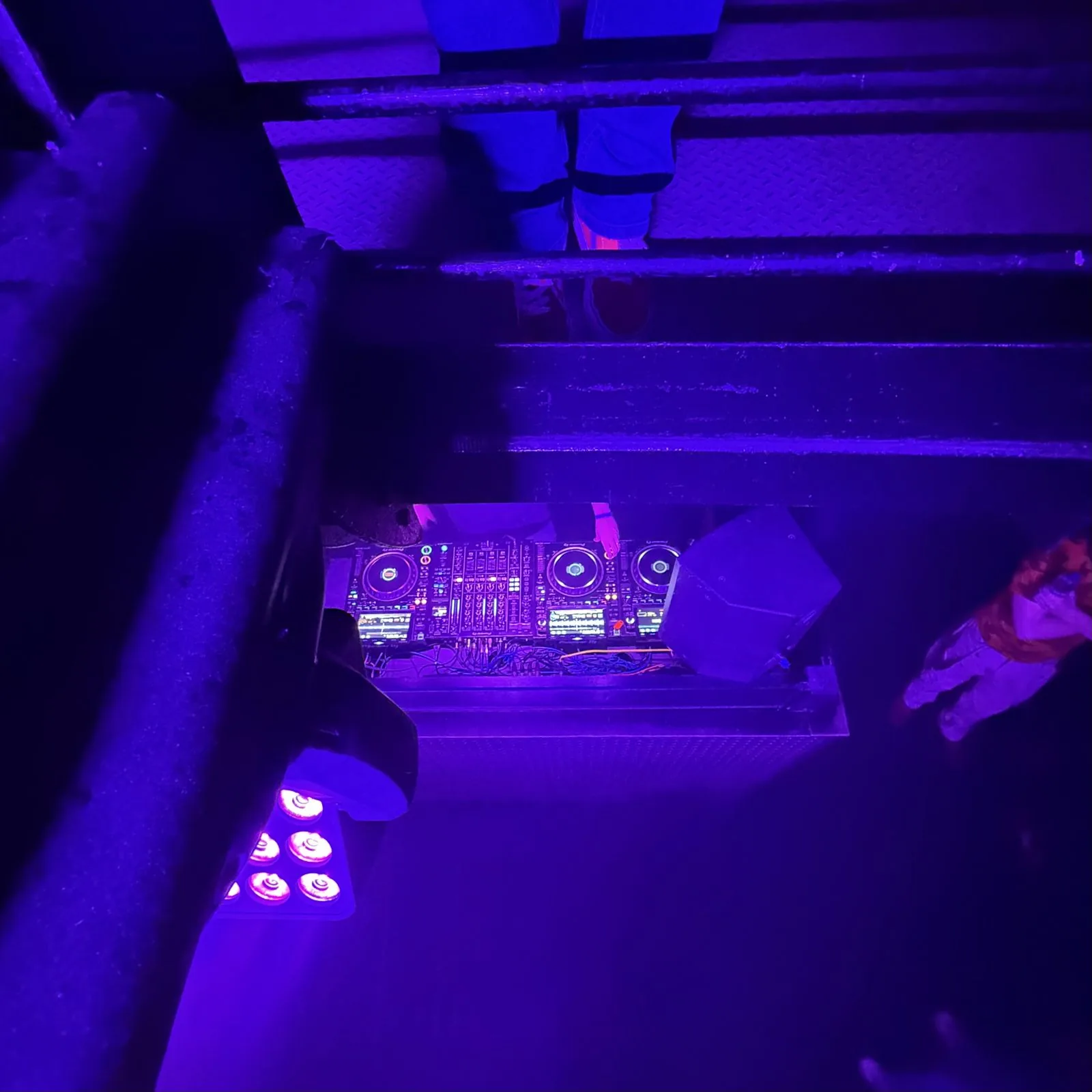 Low-angle view through scaffold rigging of a DJ booth lit in purple UV light at a live event, event photography by Hector's Eye