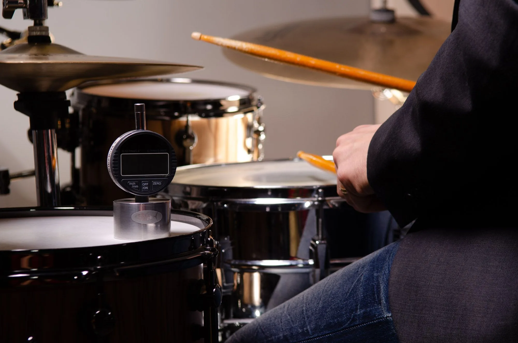 Drummer's hand holding sticks over a drum kit, with a precision drum-tuning gauge resting on a tom head in the foreground, brand photography by Hector's Eye