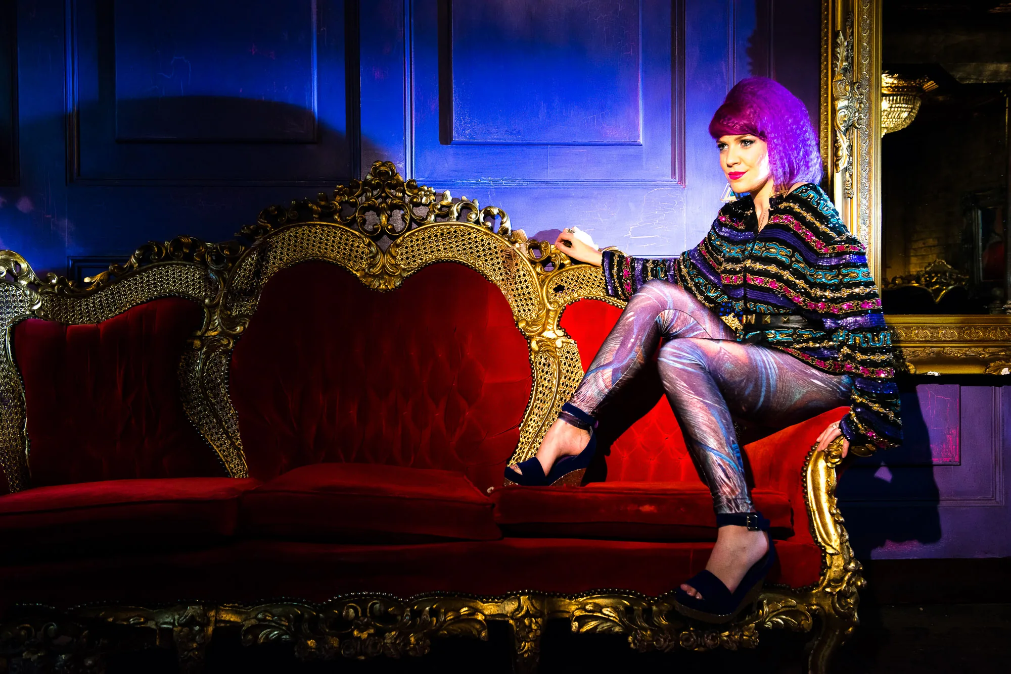 Full-length editorial portrait of a model with purple hair seated on an ornate red and gold baroque sofa, styled portrait photography by Hector's Eye