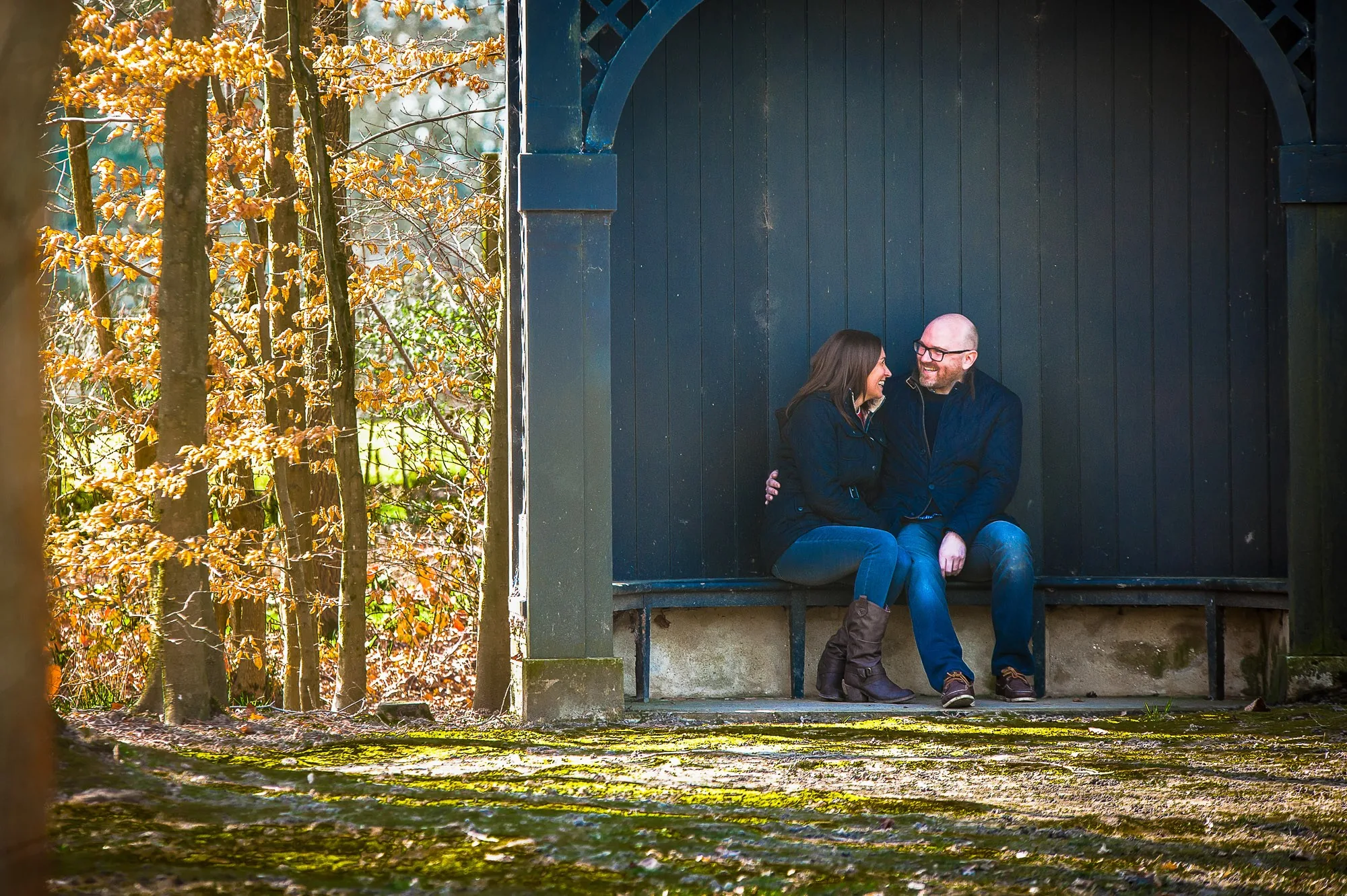 Couple laughing together in a garden shelter surrounded by autumn trees, engagement portrait photography by Hector's Eye