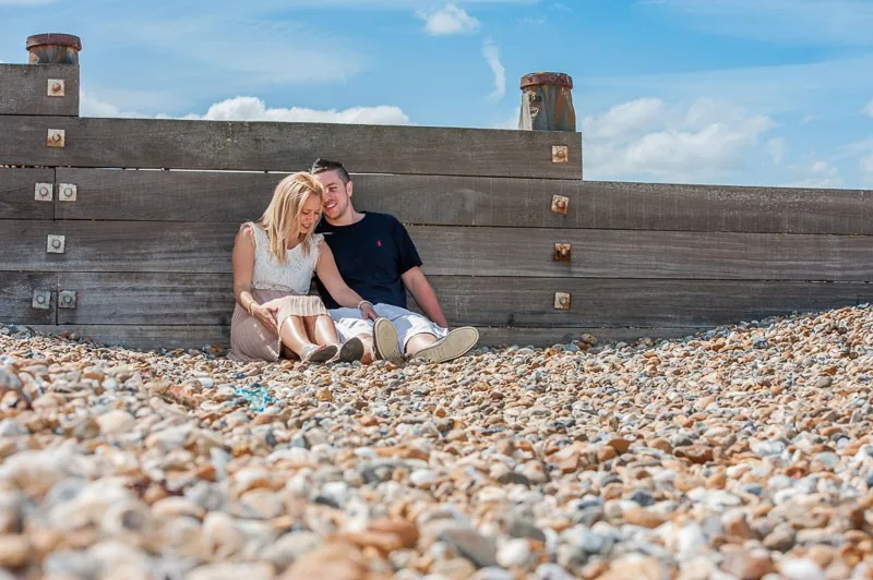 Couple sitting together on a pebble beach against a weathered wooden groyne, engagement portrait photography by Hector's Eye