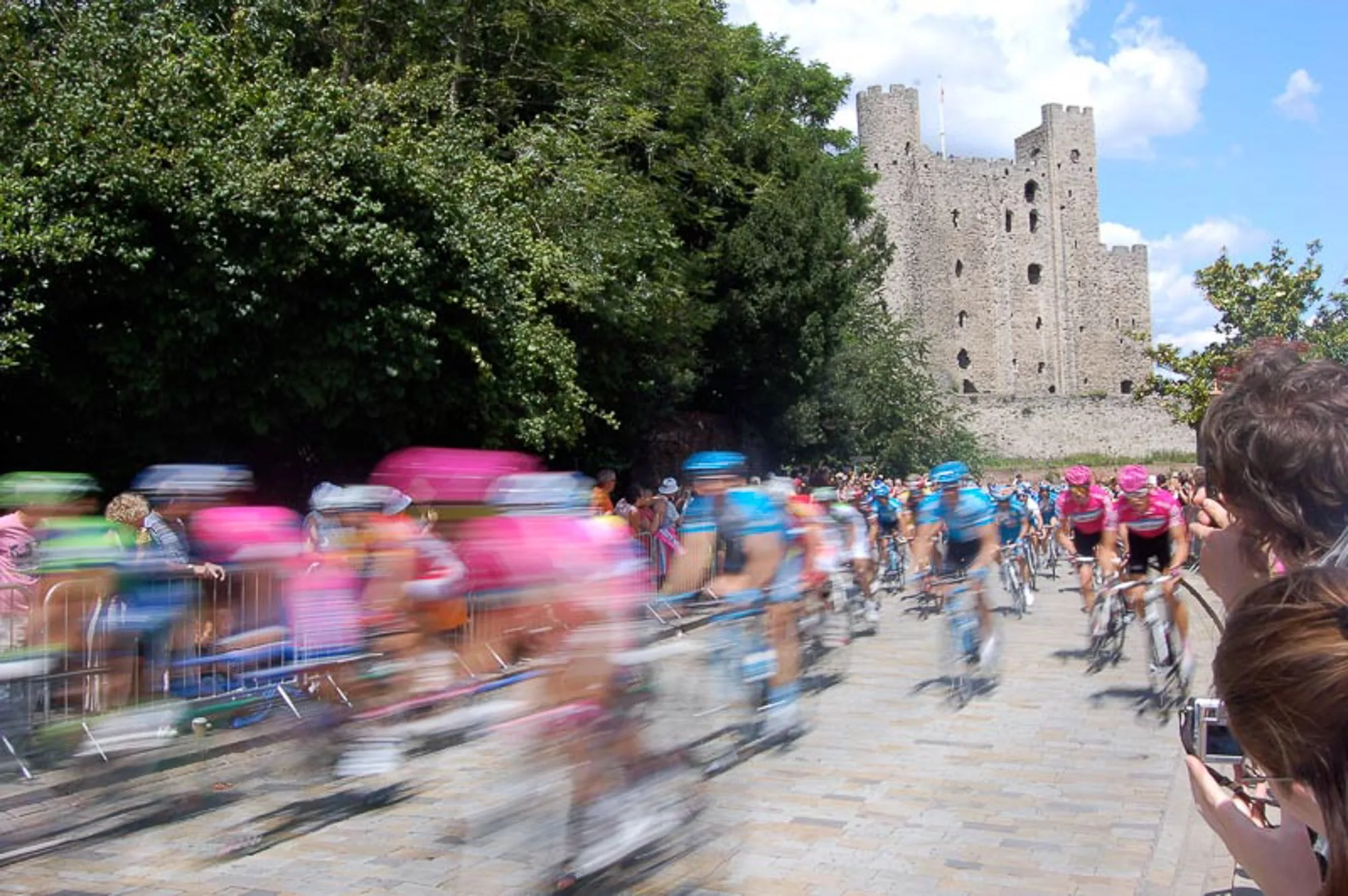 Cycling peloton racing past a historic castle gatehouse, live sports event photography by Hector's Eye
