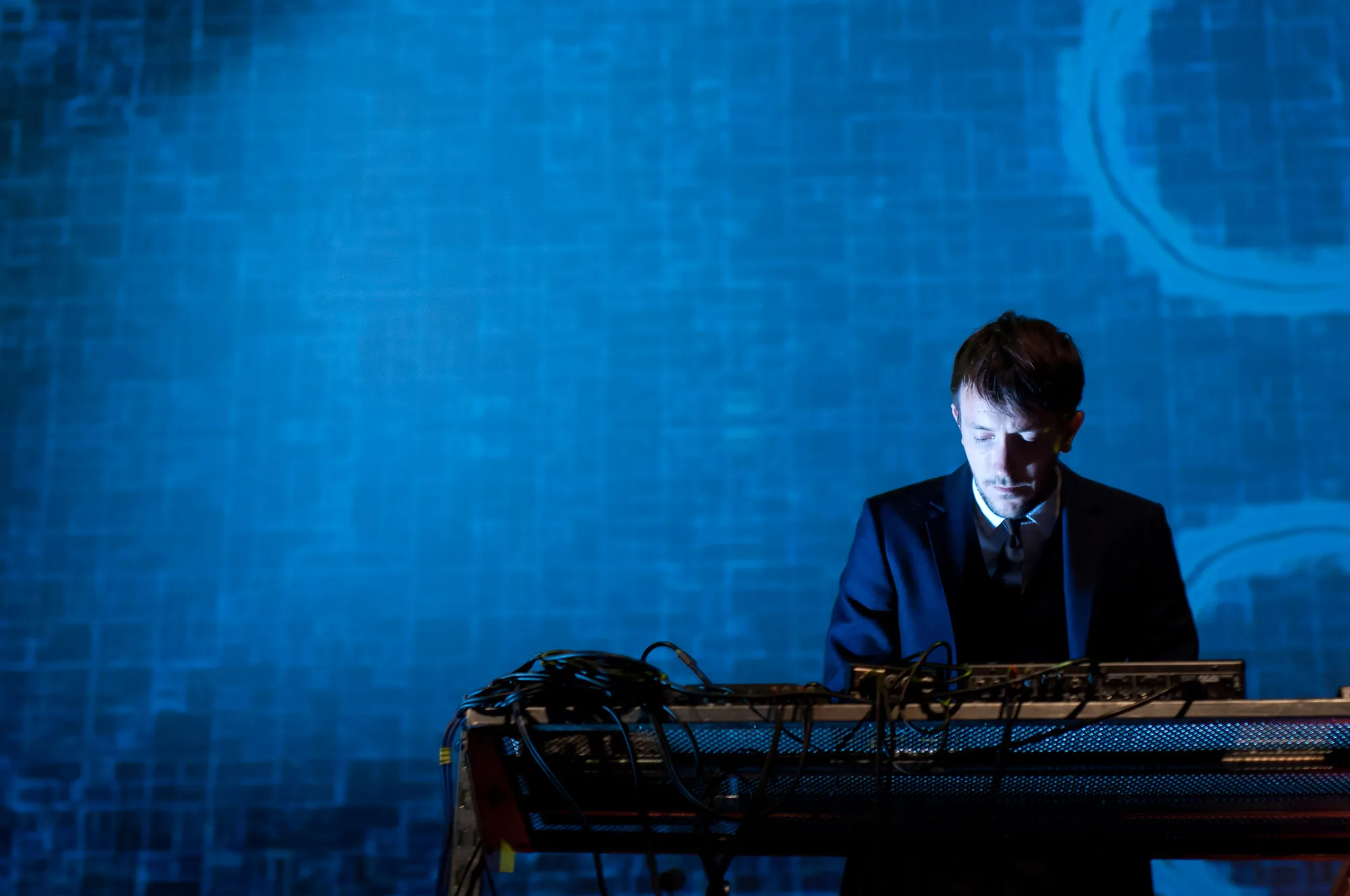 Musician performing at a keyboard rig under blue stage lighting with a digital projected backdrop, live event photography by Hector's Eye