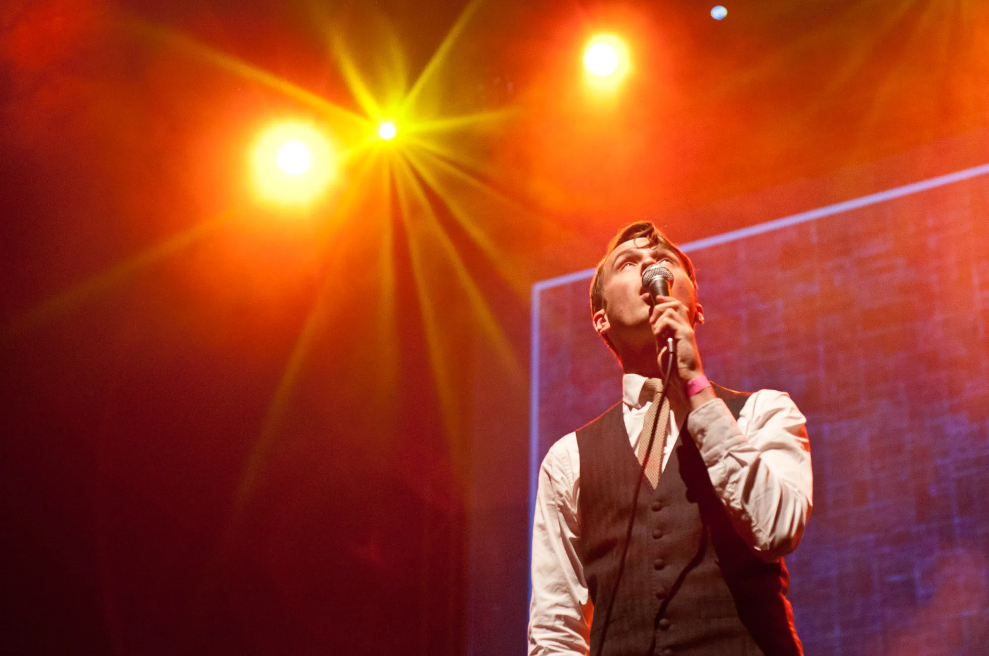 Singer performing into a microphone under warm orange starburst stage lighting, live music photography by Hector's Eye