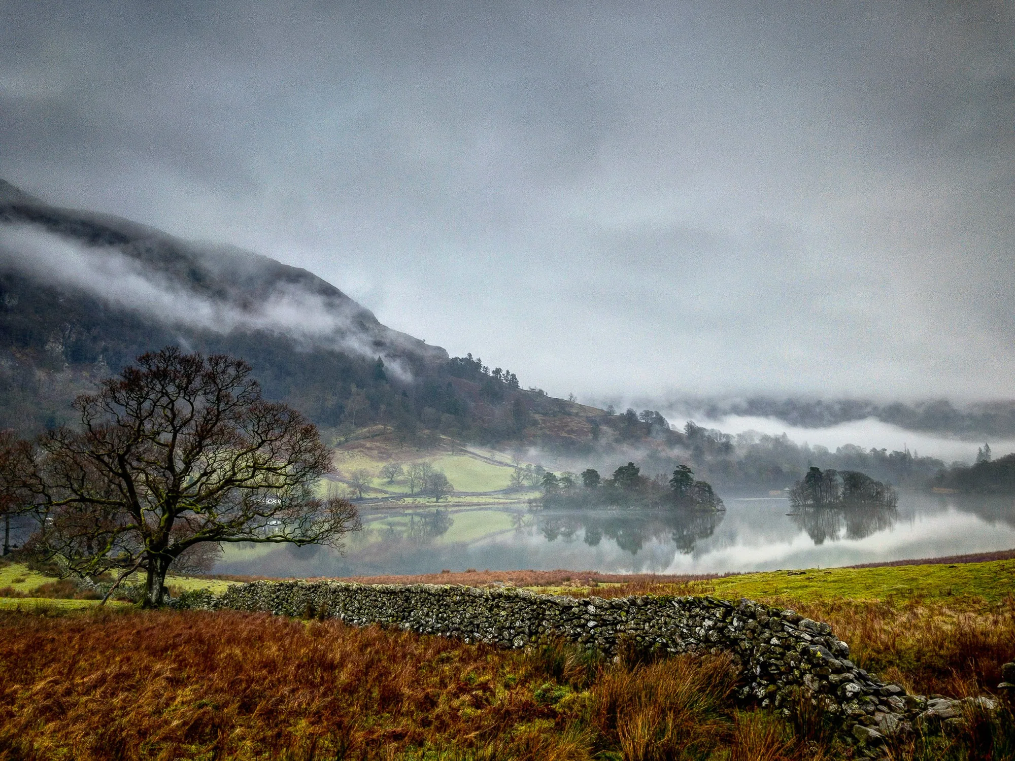 Dry stone wall beside a misty lake with cloud-covered fells behind, landscape photography by Hector's Eye