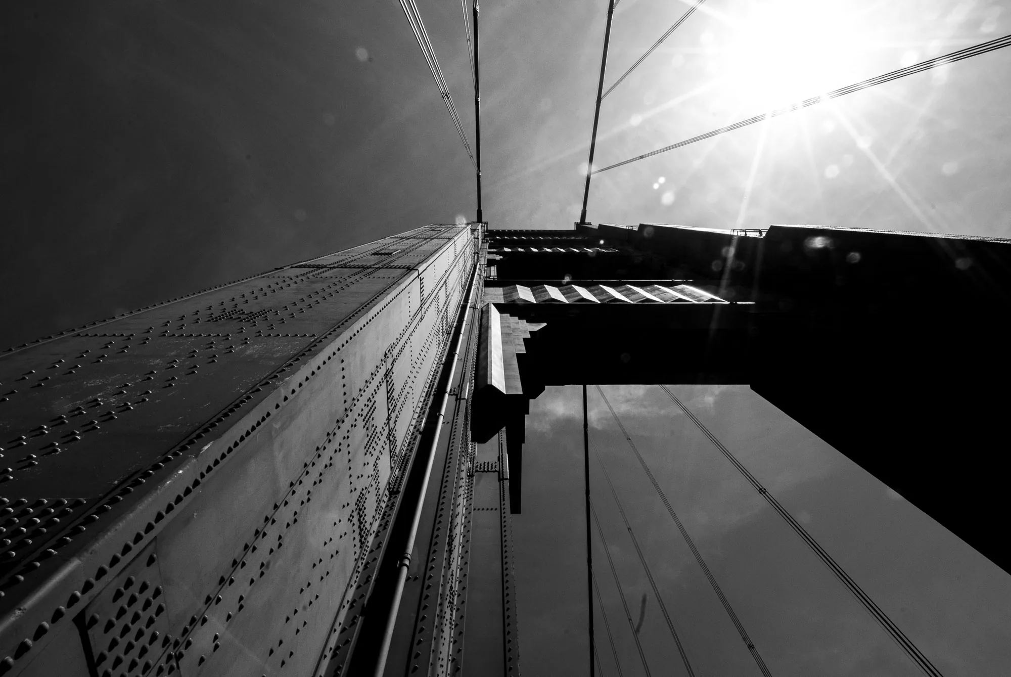Looking up a riveted suspension bridge tower into a sunburst, black and white architectural photography