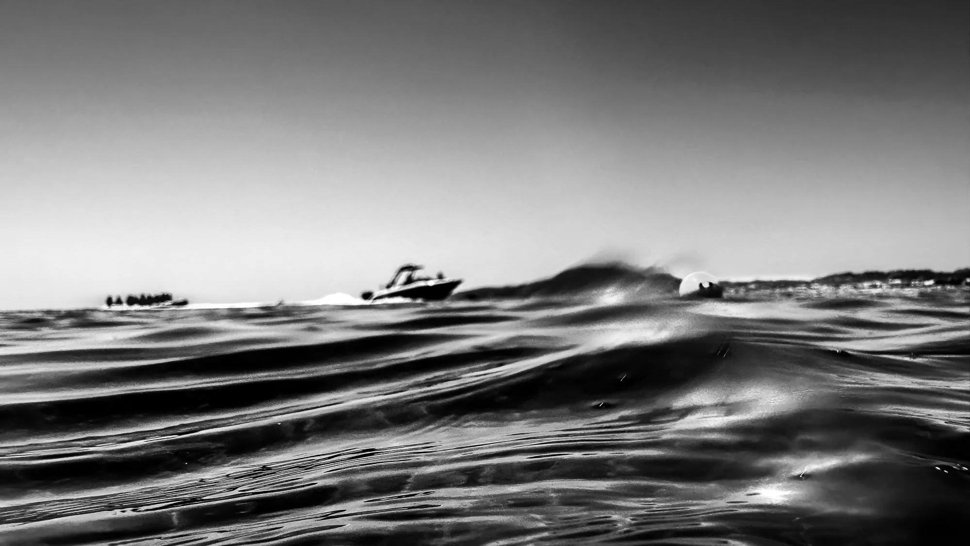 Wave breaking at water level with boats and coastline in the background, black and white abstract photography