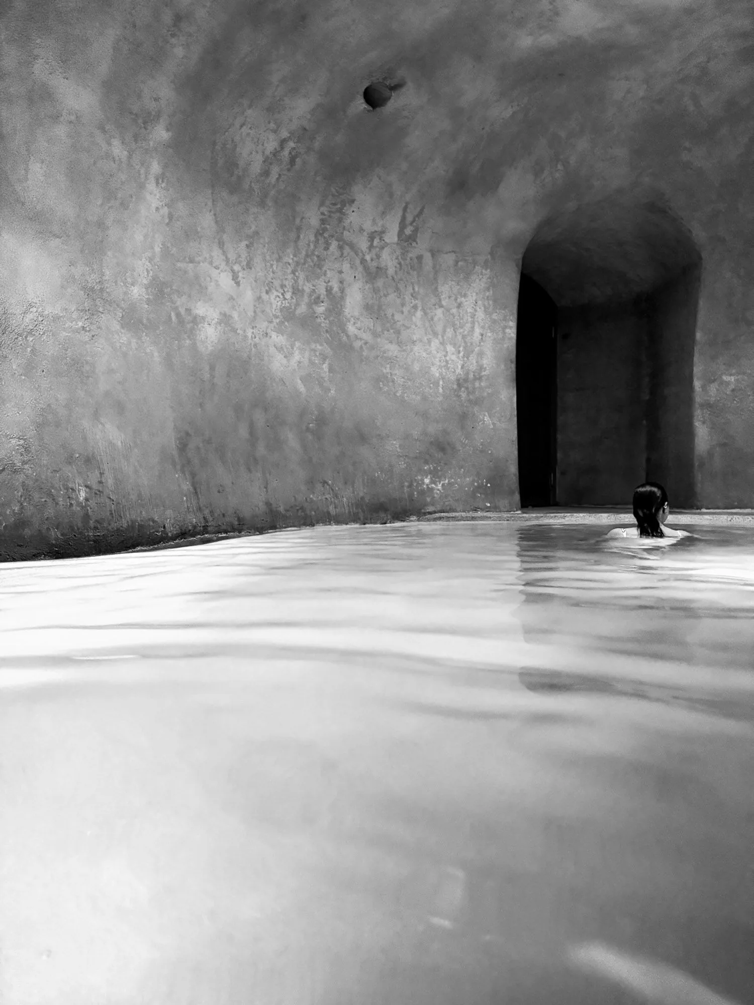 Woman swimming towards an arched doorway in a minimalist stone pool, black and white photography