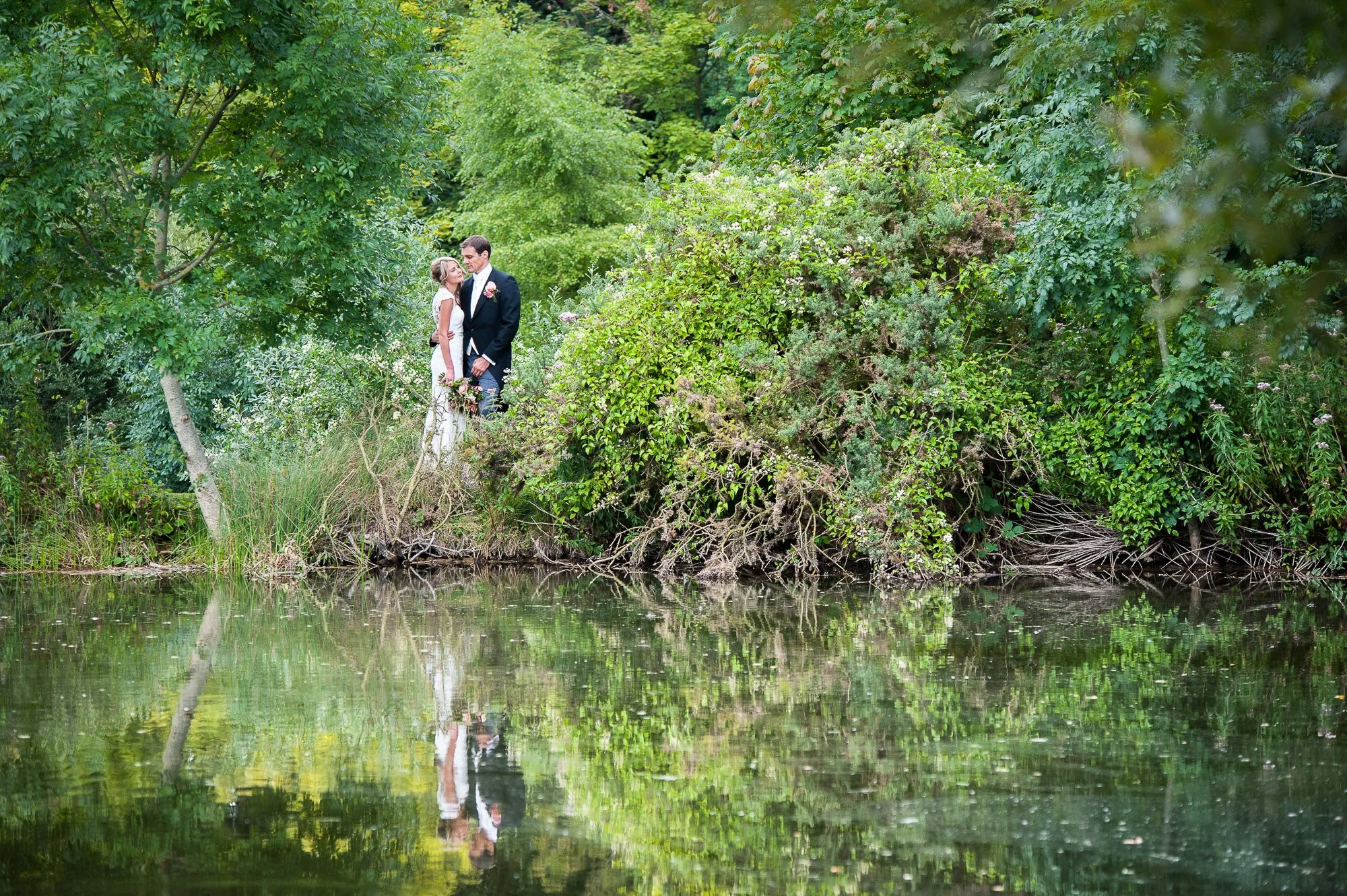 Bride and groom reflected in a still lake surrounded by woodland, wedding portrait photography by Hector's Eye